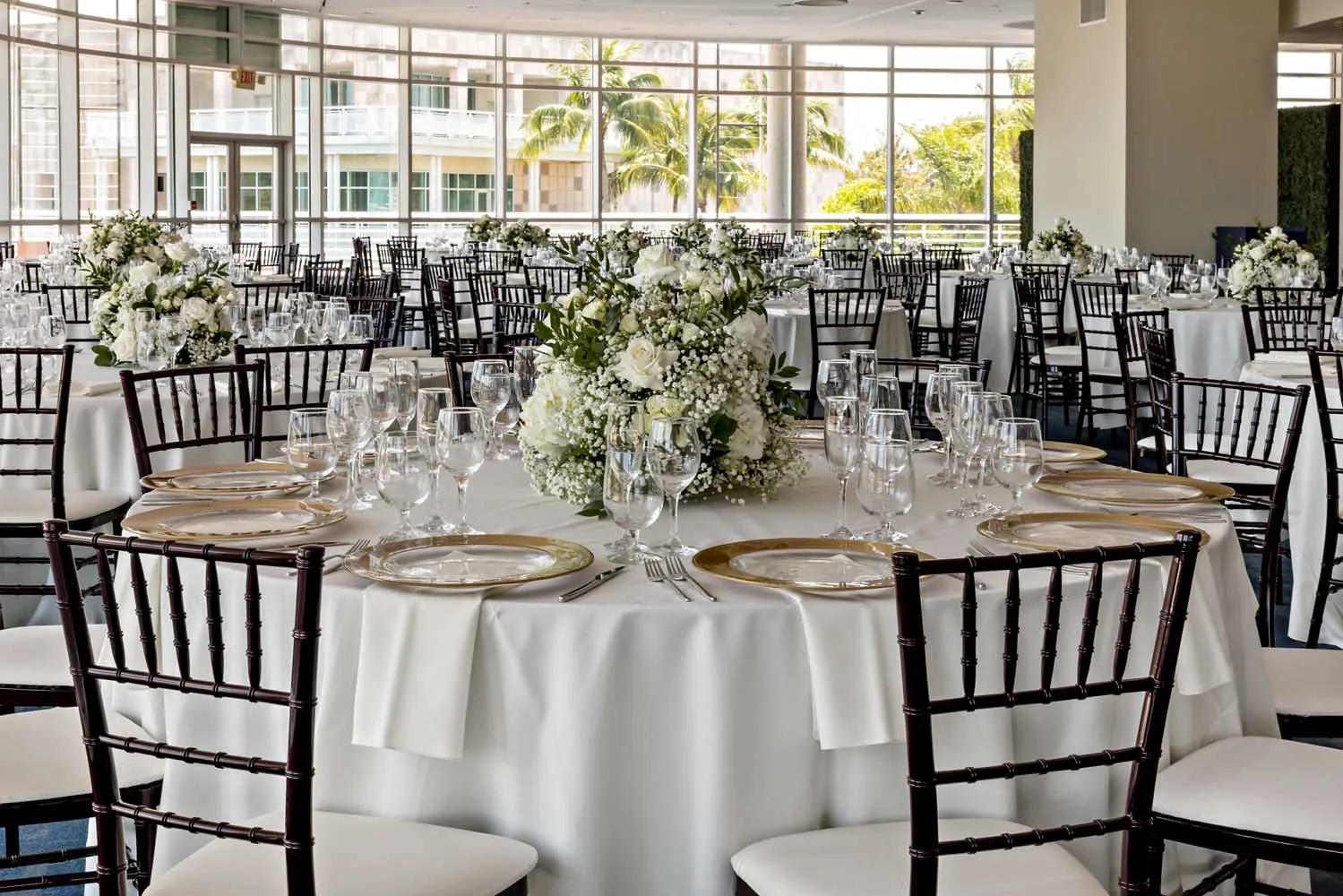 Formal reception setup in the Palm Ballroom with white florals, gold-rimmed chargers, and views of palm trees through floor-to-ceiling windows