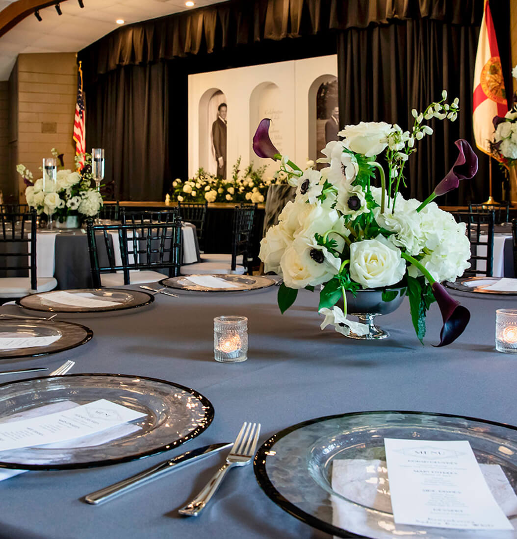 Round reception table at Venue on Lake Lily in Maitland with grey linens, white and purple floral centerpiece, and stage backdrop with portrait display