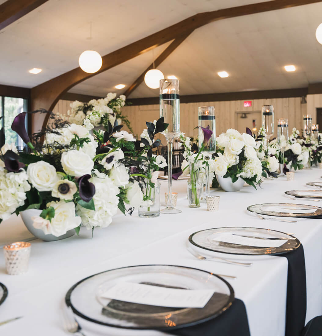 Long banquet table at Venue on Lake Lily in Maitland with white linens, purple calla lily centerpieces, floating candles, and wood-beam vaulted ceiling