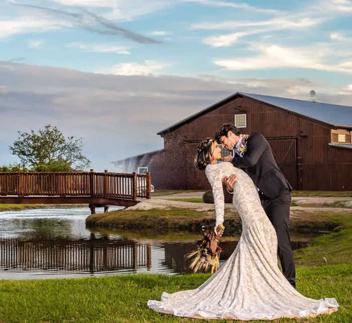 Groom lifts bride at Ever After Farms by a pond in front of a rustic white barn, with trees and floral bouquet nearby
